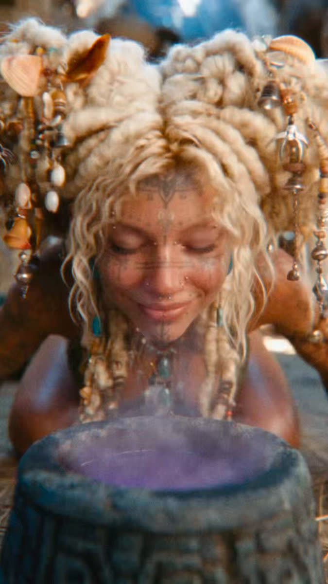 Close-up of a smiling woman with closed eyes, pale braided hair hung with shells, beads and small bells, fine tattoo lines on her face, leaning over a grey stone bowl filled with glowing purple smoke.
