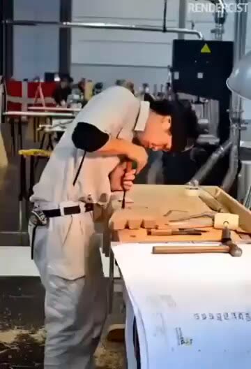 A young man in a light shirt and work trousers bends over a wooden workbench in a workshop, holding a chisel to a piece of wood, with a mallet on the bench beside him.