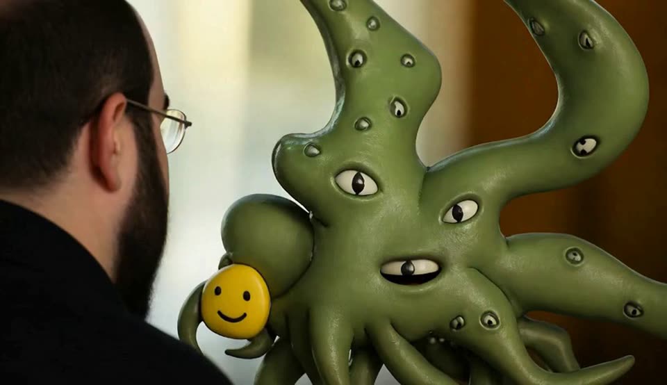 A bearded man with glasses, seen from behind, faces a green sculpted creature with many eyes and tentacles. The creature holds a small yellow smiley face against its body.