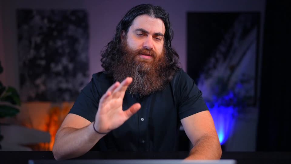 A man with long dark curly hair and a full beard, in a black short-sleeved shirt, sits at a desk and raises one open hand toward the camera. Purple and blue lighting glows behind him.
