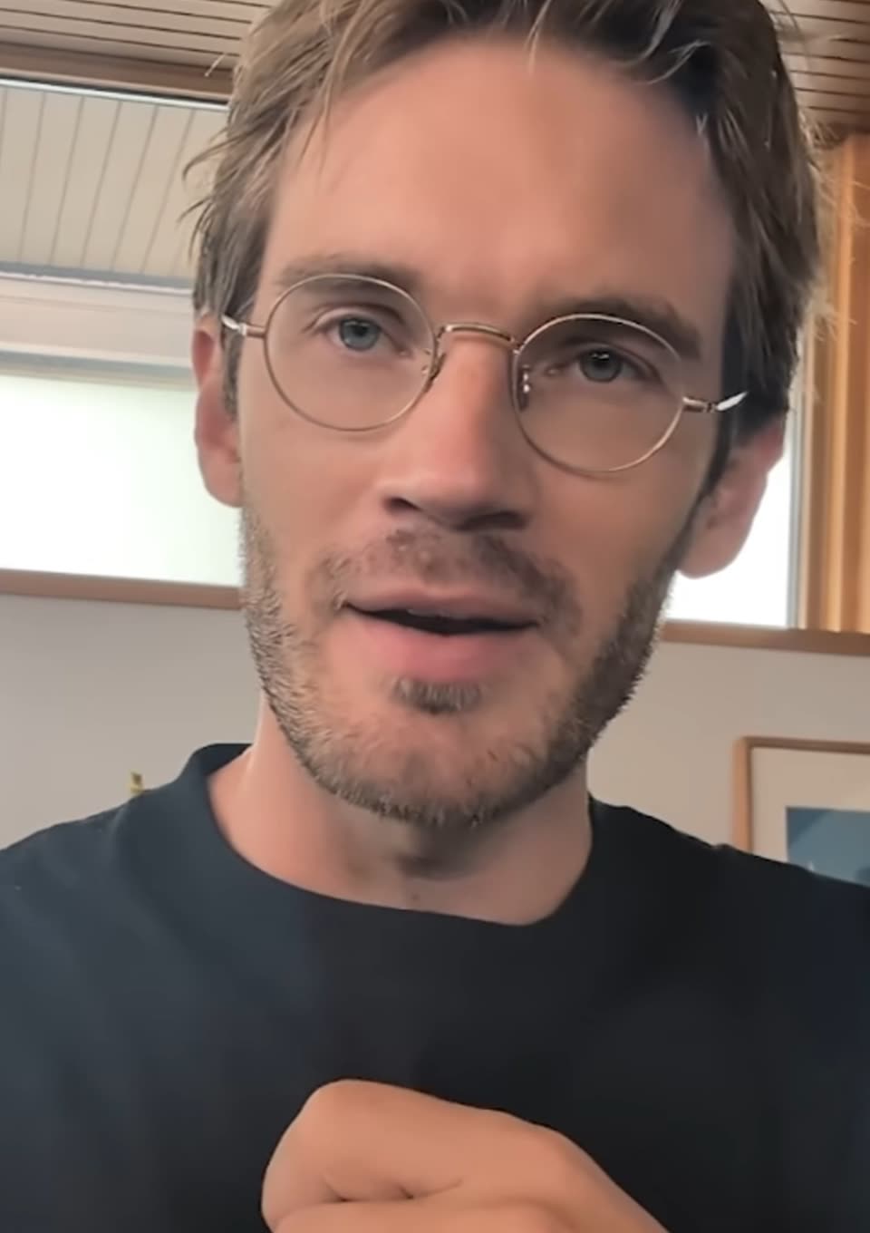 A close-up still from a video of a man with short light-brown hair, round gold-rimmed glasses and a stubbly beard, wearing a dark T-shirt and speaking to the camera in a bright room.