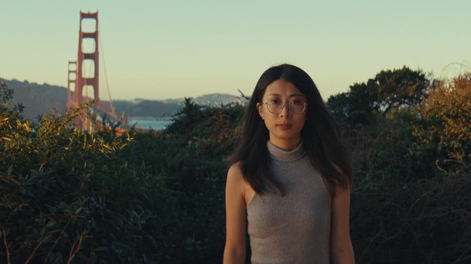 A young woman with long dark hair and round glasses, in a grey sleeveless turtleneck, stands looking at the camera among green bushes at dusk, with the Golden Gate Bridge in the distance behind her.