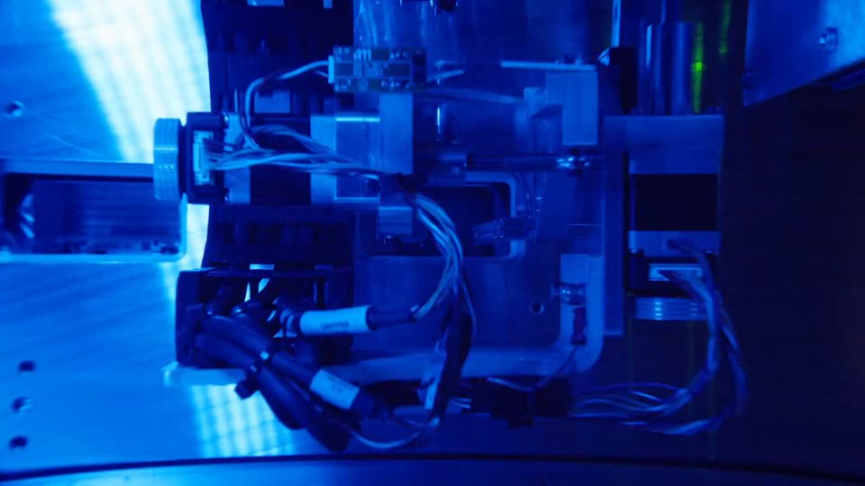 A close-up of a small machine lit in deep blue, with metal brackets, a knurled knob on the left, and bundles of grey and black wires, some with white labels.