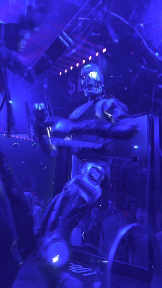 A silver humanoid robot with a metal skull face and glowing eyes, mid-motion inside a glass enclosure under blue and purple stage lights, with a crowd watching behind it.