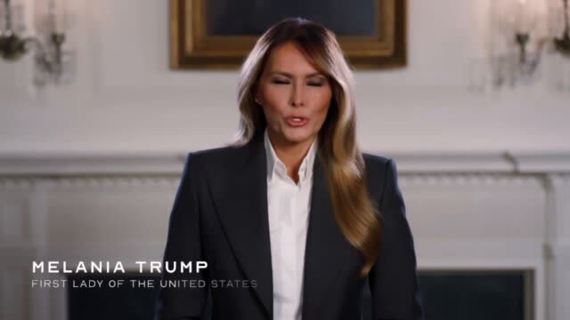Melania Trump, in a dark blazer and white shirt, speaks to camera in a formal room with a gilt-framed painting and a white mantel behind her. A caption reads Melania Trump, First Lady of the United States.