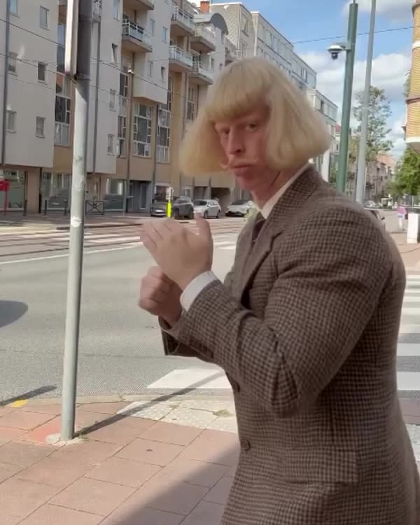 Jean Philanthrope mid-dance on a city sidewalk: a blond bowl-cut wig swinging, a thin moustache, lips pursed, fists raised in front of him, in a brown houndstooth jacket and tie.
