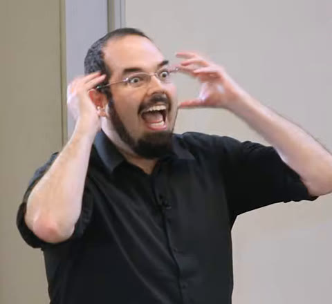 Eliezer Yudkowsky, bearded, in glasses and a black shirt, both hands raised by his head, mouth wide open in mock alarm.